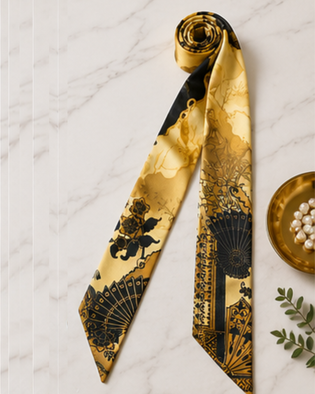 Gold and black patterned scarf on a textured white surface with a small dish of pearls and green leaves.